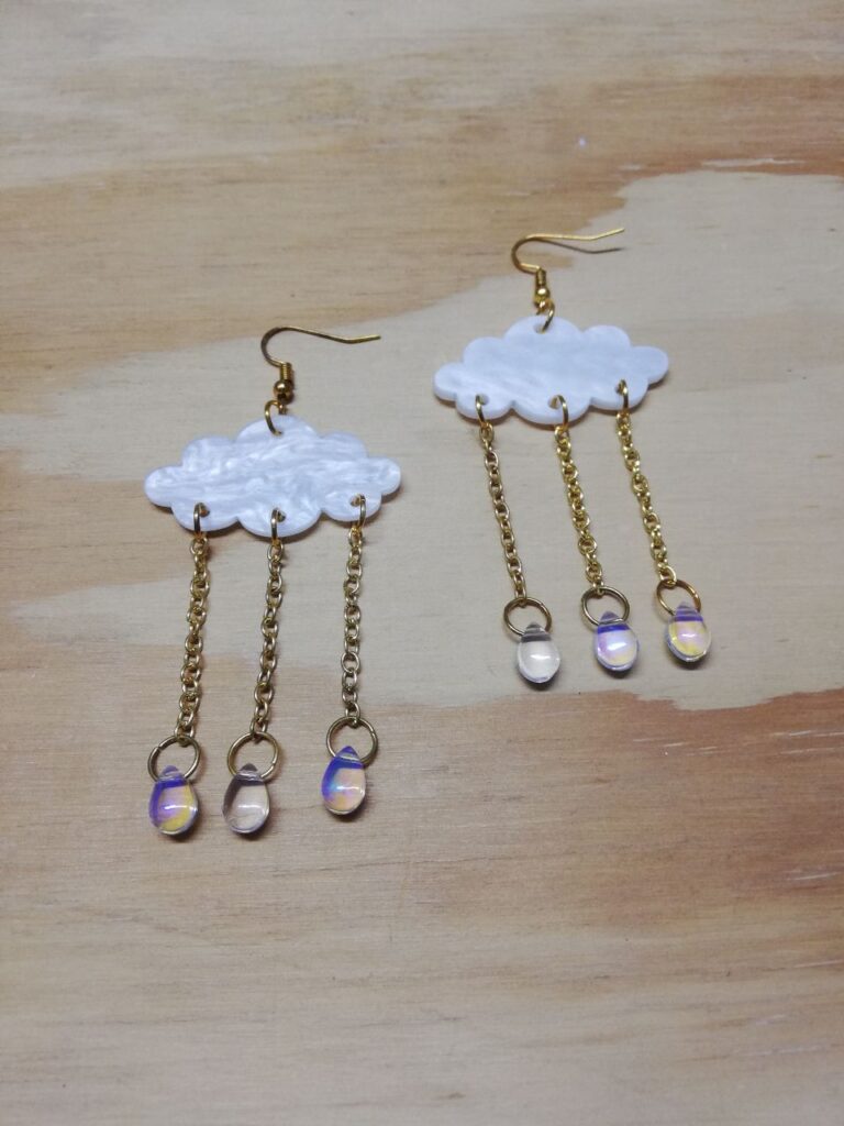 Fotografía de aretes Lluvia Iridiscente encontrados. Precio: $50 MXN Dimensiones de dije: 6.5 centímetros Material: Metal, acrílico y plástico Color de materiales: Dorado, blanco nacarado y transparente iridiscente Tipo de agarre: Gancho Categoría: Nuevo