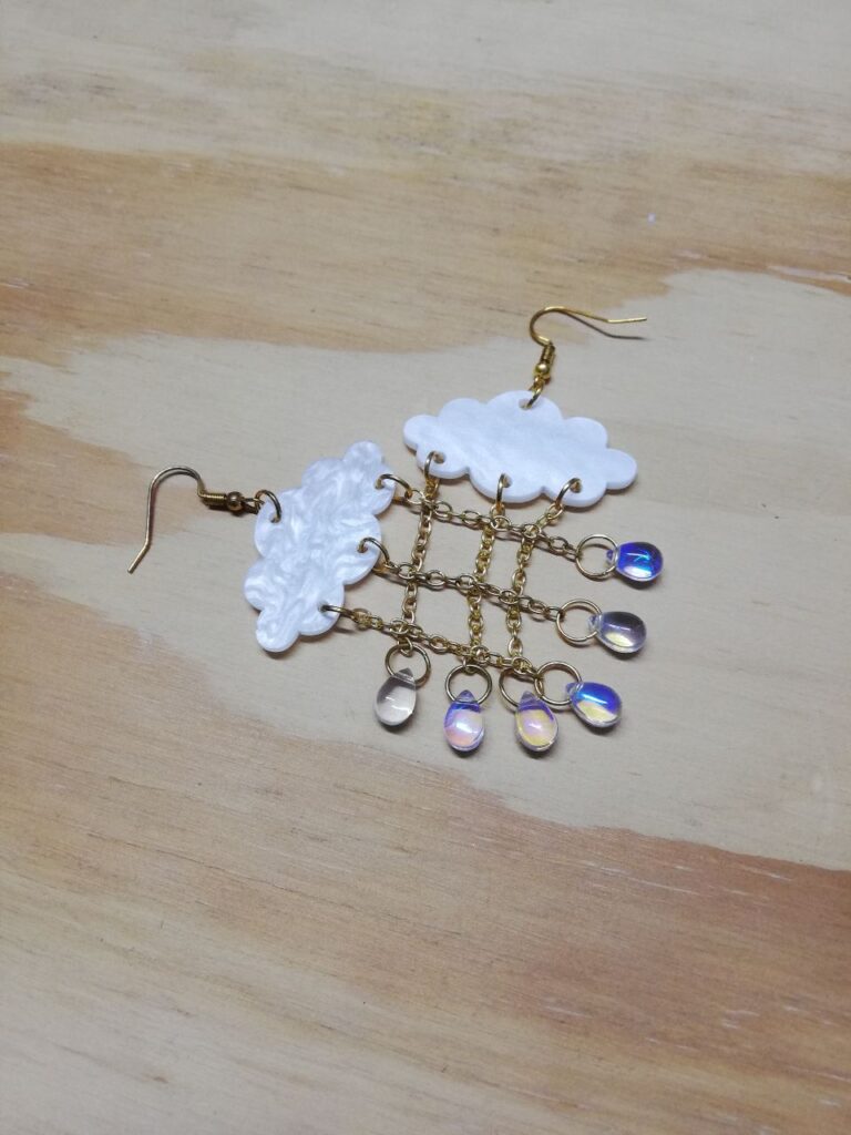 Fotografía de aretes Lluvia Iridiscente encontrados. Precio: $50 MXN Dimensiones de dije: 6.5 centímetros Material: Metal, acrílico y plástico Color de materiales: Dorado, blanco nacarado y transparente iridiscente Tipo de agarre: Gancho Categoría: Nuevo