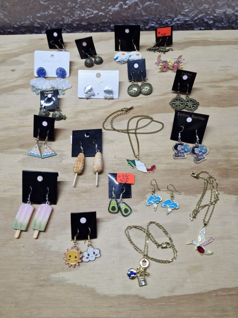 Fotografía de una selección de aretes y dijes con cadena de Almacenes Heródoto sobre mesa de madera. Se ven aretes de lluvia, hemisferio, ajolote, rosa de los vientos, mar y luna, Mafalda geógrafa, México, elote preparado, paleta de hielo, sol y nube, colibrí, aguacate, y mundo con pasaporte.