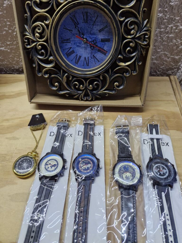 Fotografía de distintos modelos de relojes disponibles en Almacenes Heródoto desplegados sobre mesa de madera y pared de tirol color lila. De arriba a abajo y de izquierda a derecha se ven: Reloj de pared: $125 MXN. Reloj de bolsillo dorado con cadena y carátula de rosa de los vientos: $250 MXN. Reloj de pulsera grande con correa de plástico negra y vivo gris: $125 MXN. Reloj de pulsera grande con correa de plástico negra y vivo marrón: $125 MXN. Reloj de pulsera grande con correa de imitación de piel negra: $125 MXN. Reloj de pulsera grande con correa de plástico negra y vivo blanco: $125 MXN.