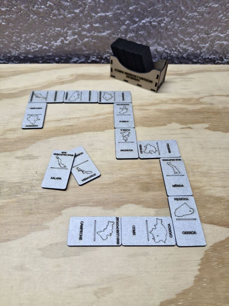 Fotografía de dominó de estados y capitales de México con un ejemplo de uso. Precio: $50 MXN Dimensiones: Caja llena: 6 cm. por 9 cm. por 3.8 cm.; Ficha: 0.25 cm. por 3 cm. por 5.5 cm. Color: Frente: Plateado; Detrás: Marrón oscuro Material: Madera industrializada Grabado y cortado con láser Idioma: Español Categoría: Nuevo