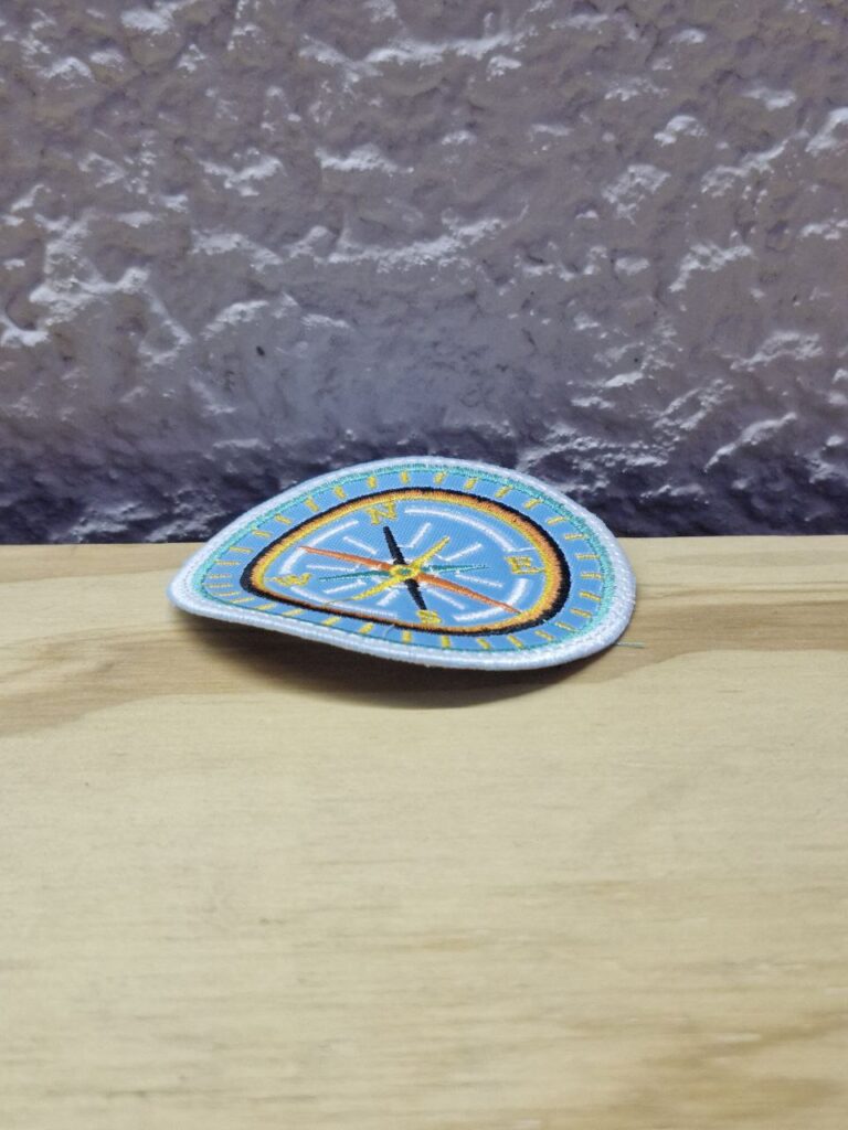 Fotografía de aplicación bordad de brújula. Precio: $30 MXN Dimensiones: Diámetro 7.3 cm. Material: Textil. Colores: Celeste con vivos en amarillo, negro, amarillo y blanco. Categoría: Nuevo.