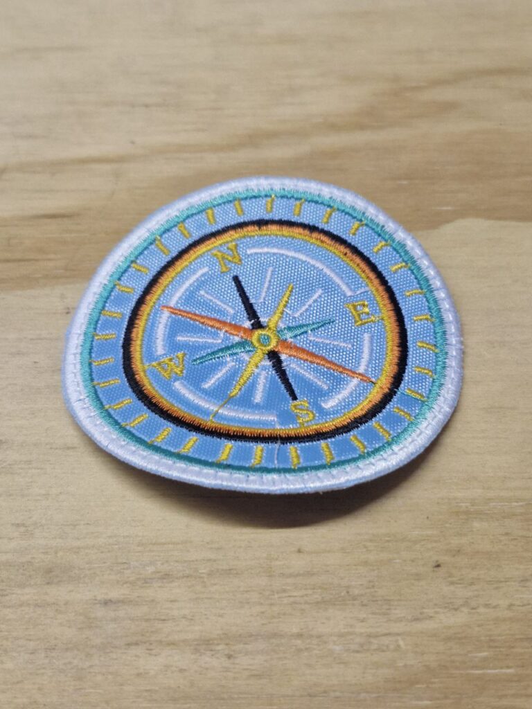 Fotografía de aplicación bordad de brújula. Precio: $30 MXN Dimensiones: Diámetro 7.3 cm. Material: Textil. Colores: Celeste con vivos en amarillo, negro, amarillo y blanco. Categoría: Nuevo.