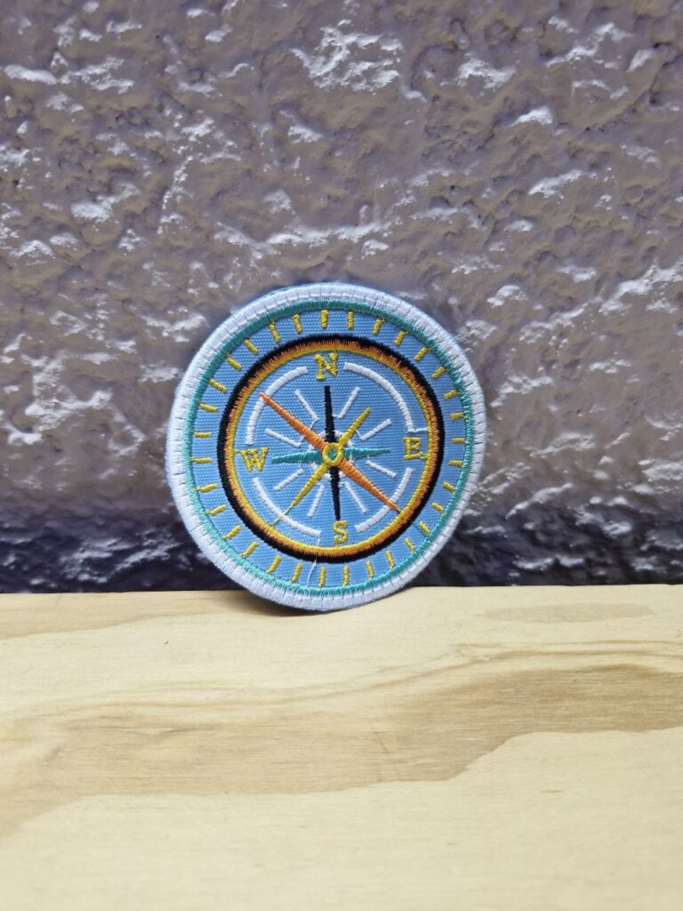Fotografía de aplicación bordad de brújula. Precio: $30 MXN Dimensiones: Diámetro 7.3 cm. Material: Textil. Colores: Celeste con vivos en amarillo, negro, amarillo y blanco. Categoría: Nuevo.