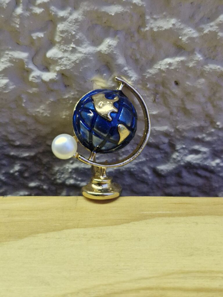 Fotografía de frente de broche Globo Terráqueo y Luna. Precio: $100 MXN. Dimensiones aproximadas: Ancho 3 cm. x Alto 3.6 cm. x Grueso 1.8 cm Material: Metal y plástico. Colores: Dorado, azul y aperlado. Categoría: Nuevo.