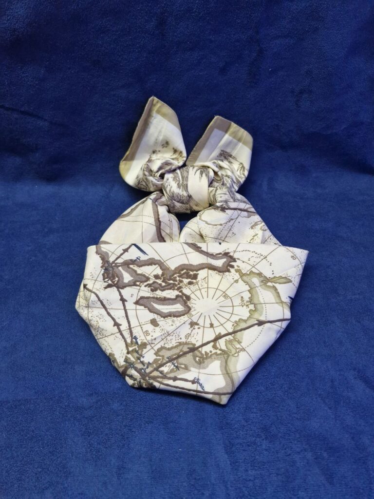 Fotografía de mascada Around the World anudada sobre mesa de terciopelo marino. Precio: $100 MXN Talla: Unitalla Dimensiones: 90 cm. x 90 cm. Material: 100% Poliéster tacto sedoso. Colores: Marrones y beige. Categoría: Nuevo